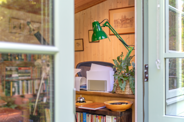 Inside a Langham Studio home study View through open double doors into a timber-lined garden office with a desk, green task lamp, books and potted plant.